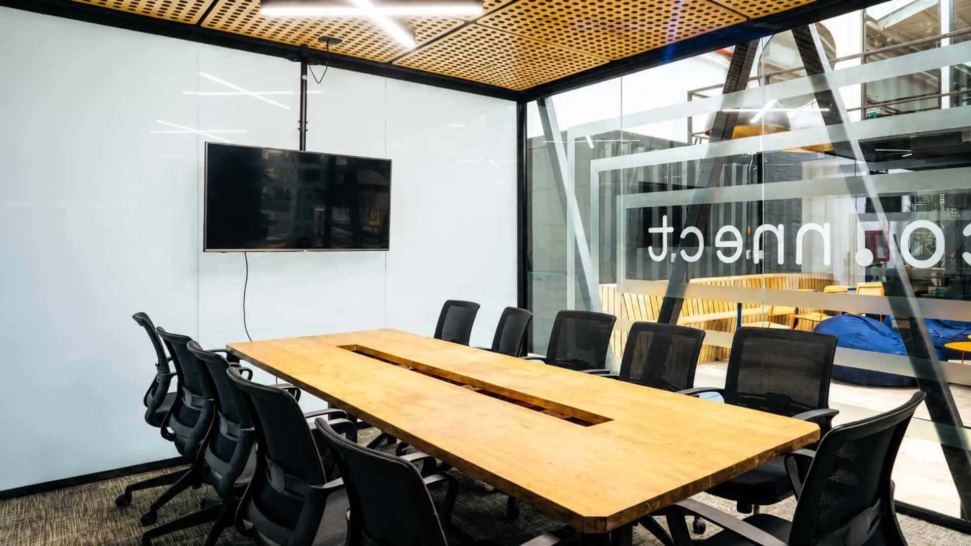 NOI meeting room in Medellín with a long wooden table, black ergonomic chairs, a wall-mounted TV and a glass partition facing the lounge